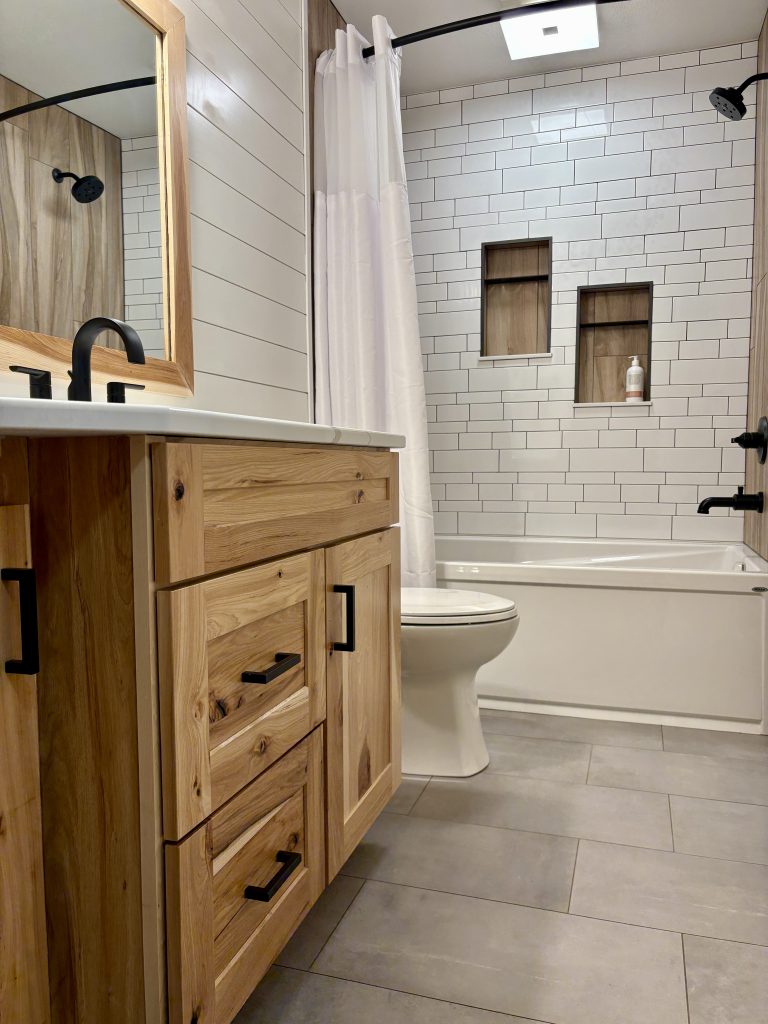 Modern hallway bathroom remodel in Northwest Ohio featuring custom maple vanity, quartz countertop, matte black fixtures, subway tile tub surround, and porcelain tile flooring.
